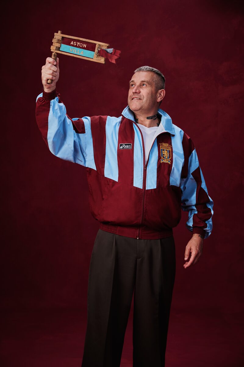 Man wearing a claret and blue Aston Villa Asics jacket holding up a handmade wooden stop-motion animation sign reading "Aston Villa" against a dark red background